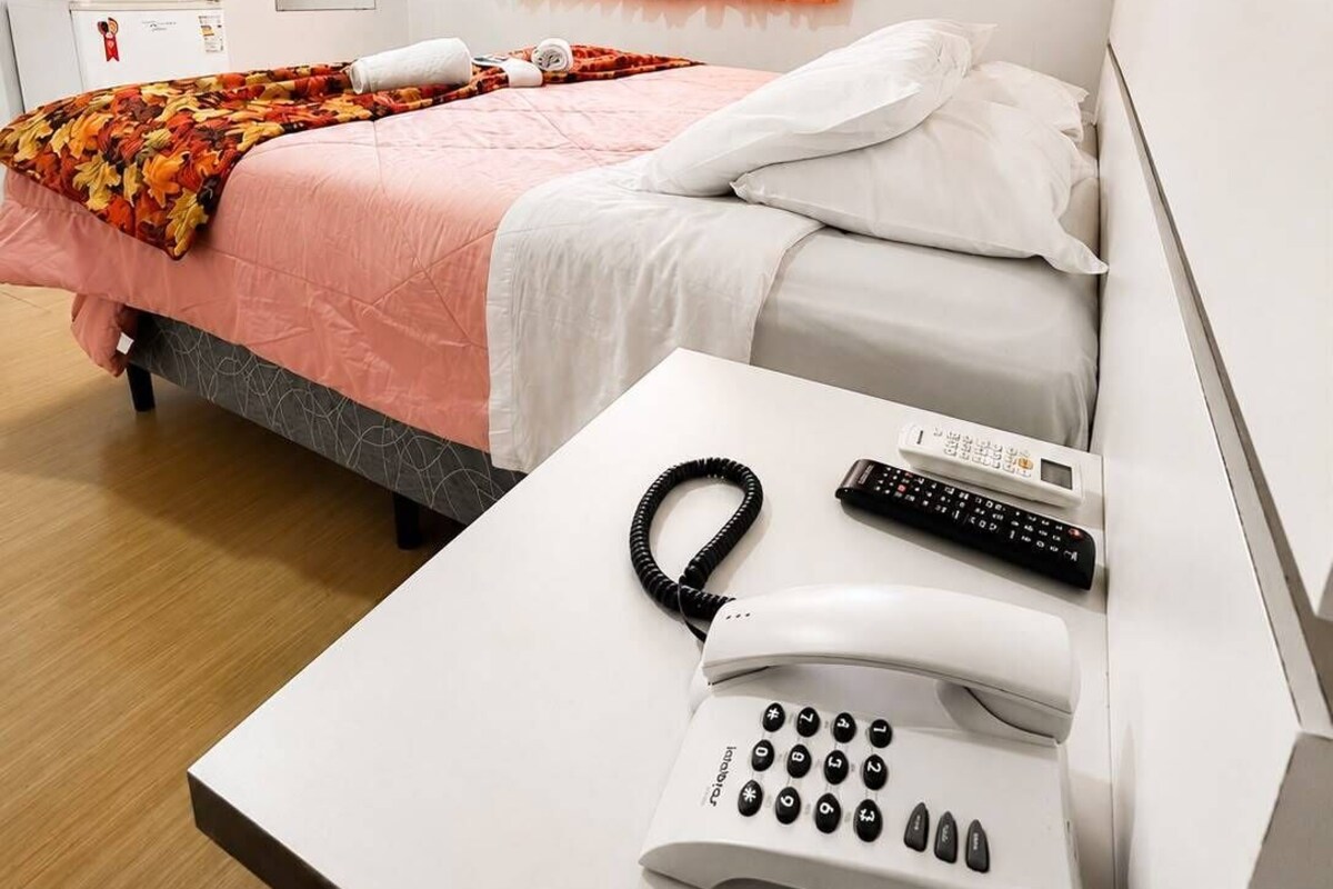 A hotel room with a bed, a bedside table with a phone and remote, a TV mounted on the wall, and an air conditioning unit.