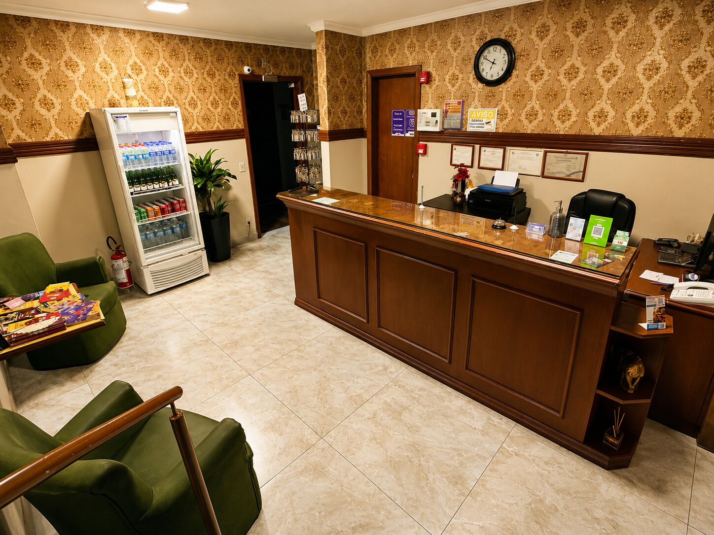 A hotel reception area with a reception desk, a refrigerator stocked with beverages, a clock on the wall, and a potted plant.