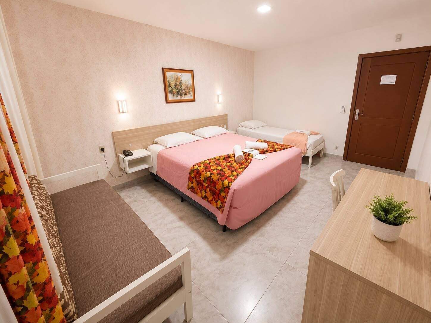 A hotel room with a bed, a desk, a chair, a bedside table, and a door.