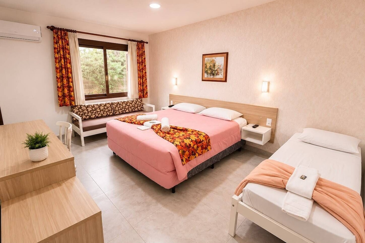 A hotel room with a bed, a desk, a chair, a window with curtains, a bedside table, and a wall-mounted picture.