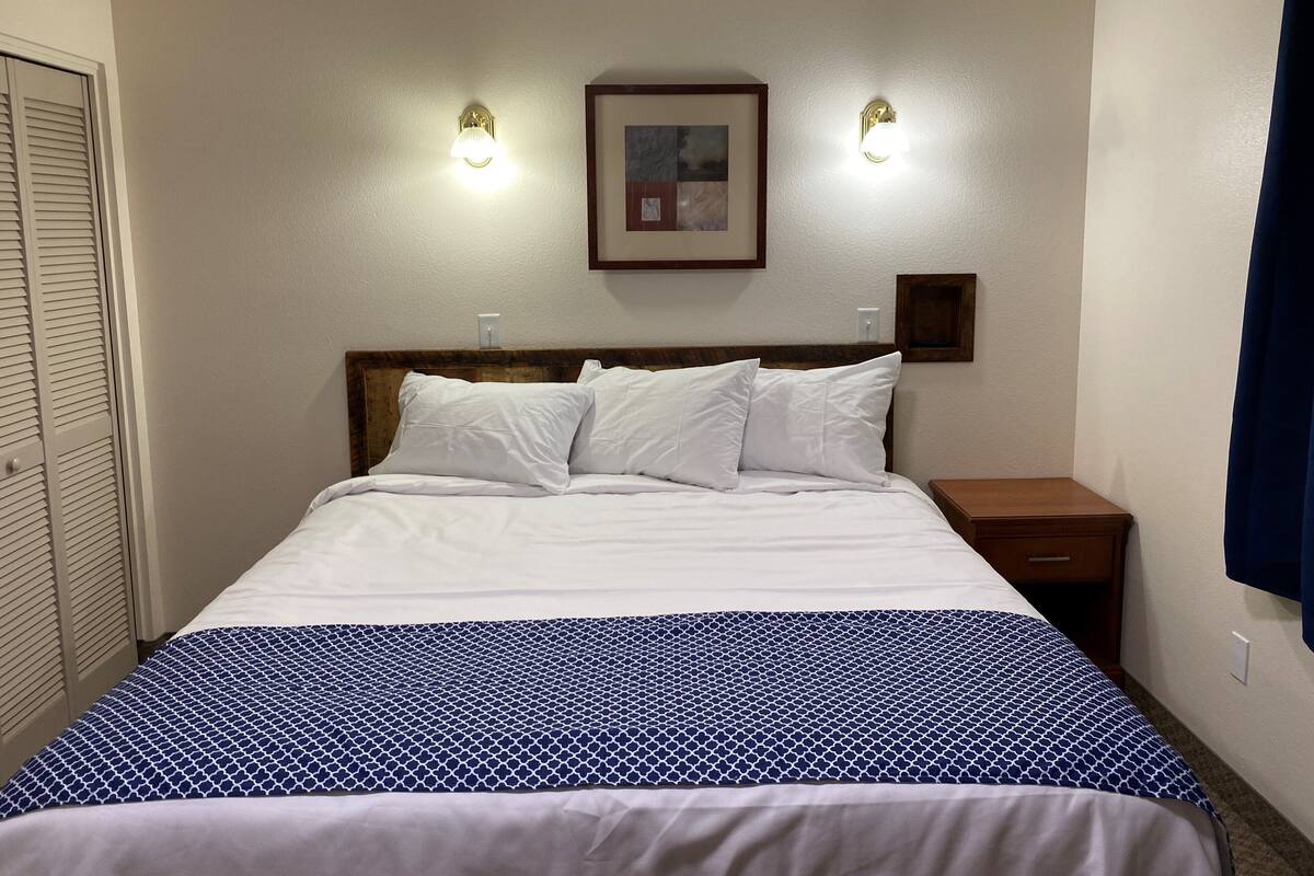A neatly made bed with a patterned bedspread, two wall-mounted lights, a framed picture, and a wooden nightstand.