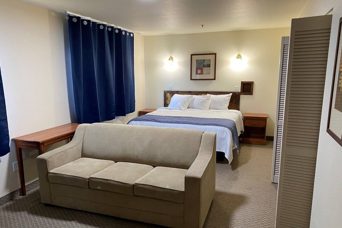 A hotel room with a beige sofa, a bed with a blue and white patterned bedspread, a wooden nightstand, and a framed picture on the wall.