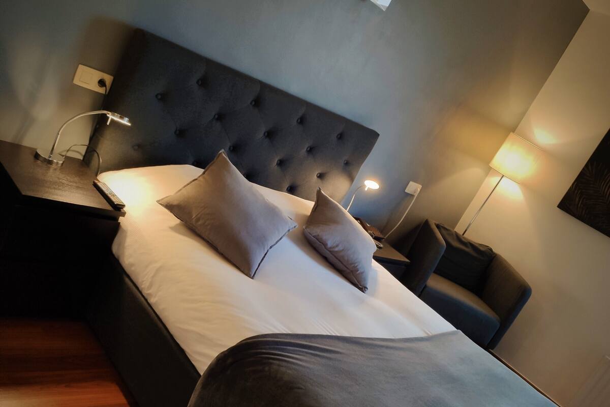 A neatly made bed with a grey blanket, two pillows, and a headboard. There are two bedside lamps and a small table with a remote control.