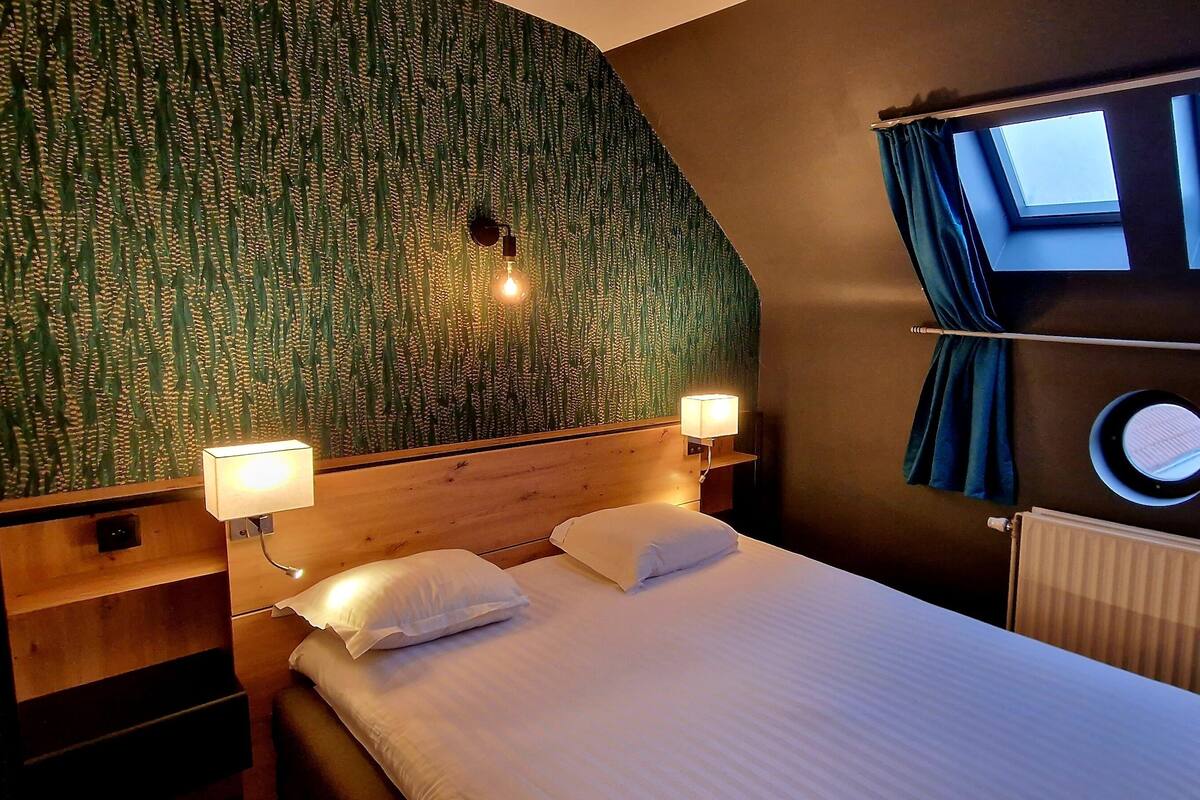 A hotel room with a bed, two bedside lamps, a headboard, a window with curtains, and a wall with a textured pattern.