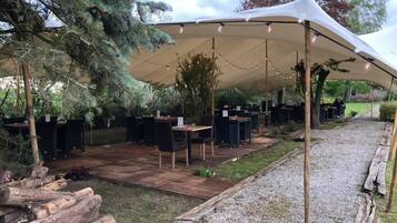 Un área de comedor al aire libre con mesas y sillas bajo una gran carpa, rodeada de follaje y árboles.