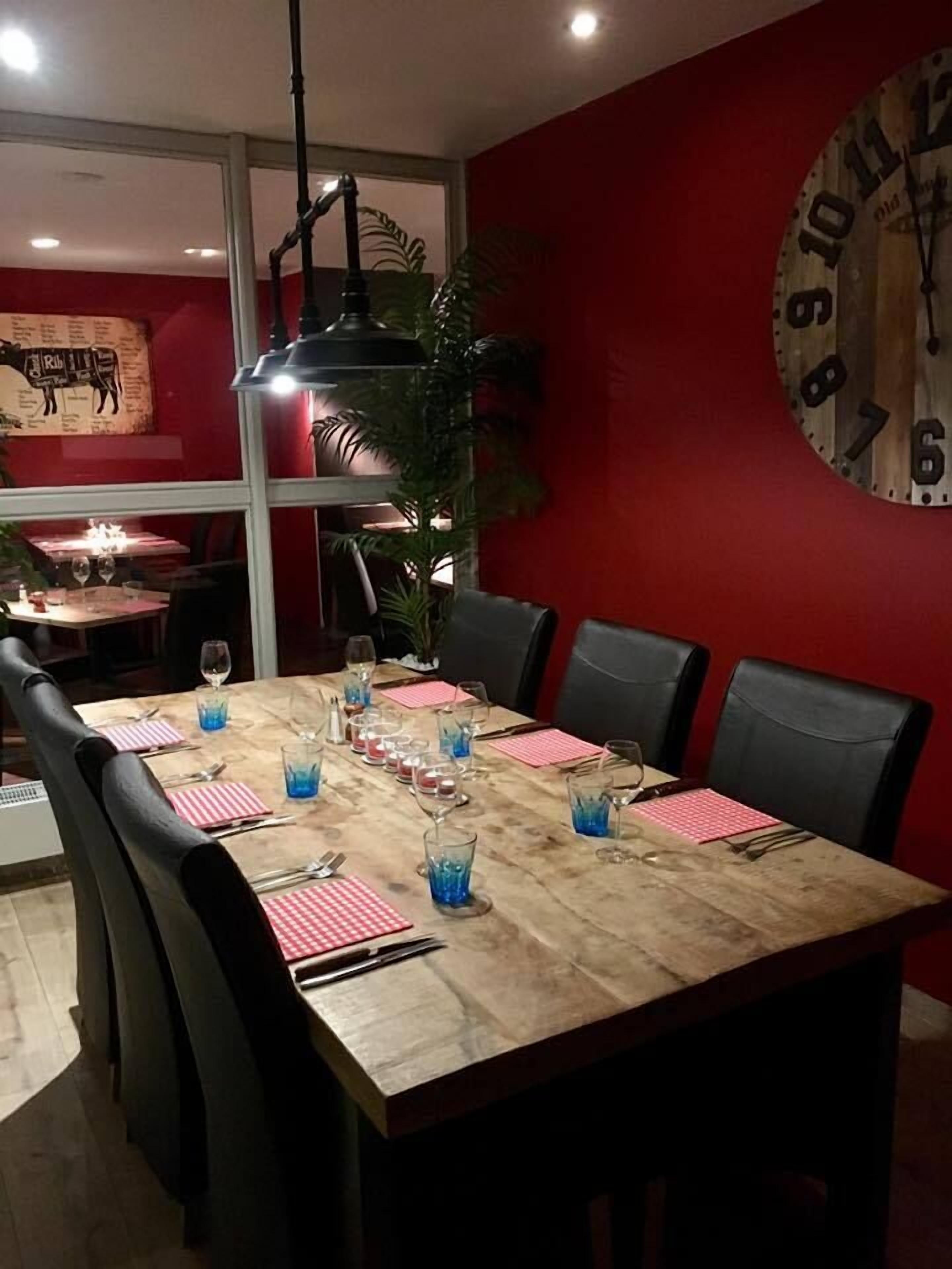A dining area with a wooden table set for six, surrounded by black chairs. A large clock is mounted on the red wall.