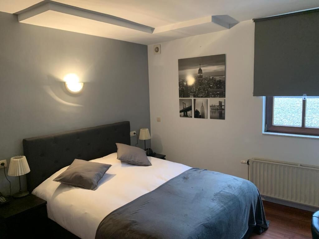 Une chambre à coucher avec un lit, deux lampes de chevet, un luminaire fixé au mur, une fenêtre donnant sur un beau paysage et un tableau représentant une vue de la ville accroché au mur.