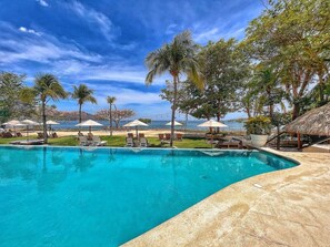 A swimming pool with lounge chairs and umbrellas, surrounded by palm trees and a view of the ocean.
