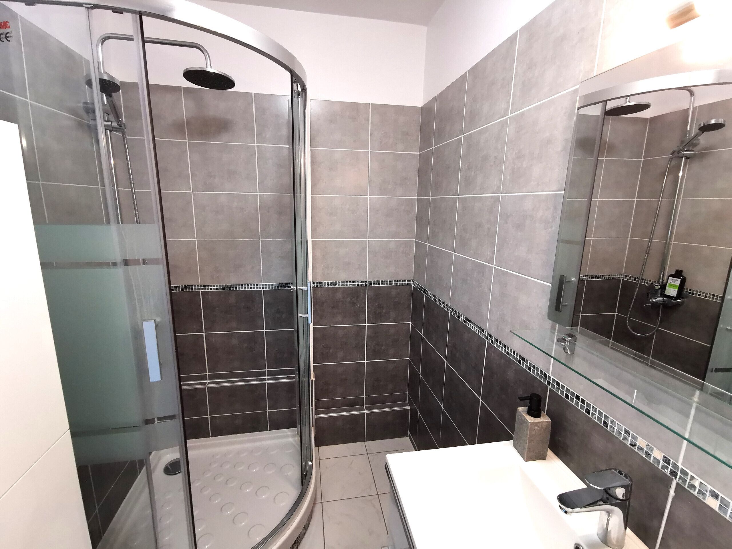 Une salle de bain moderne dotée d’une douche à parois de verre, de murs recouverts de carreaux foncés et d’un lavabo blanc avec un plan de travail en verre.