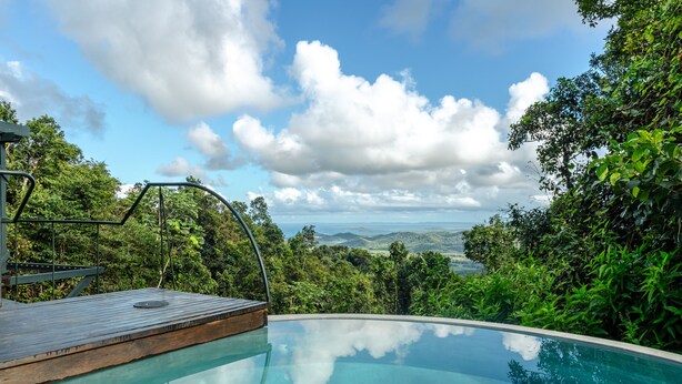 Una piscina infinita privada con cubierta de madera y barandillas metálicas, rodeada de exuberante vegetación y una vista panorámica de colinas distantes.
