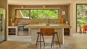Una cocina moderna con una isla grande, taburetes de bar, un refrigerador y una ventana con vista a follaje.