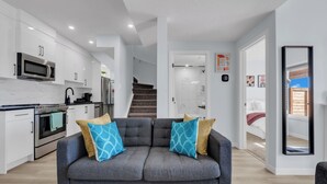 Una cocina moderna con armarios blancos, un microondas y una estufa. En la sala de estar, hay un sofá gris con cojines estampados. Una escalera conduce a otra habitación.