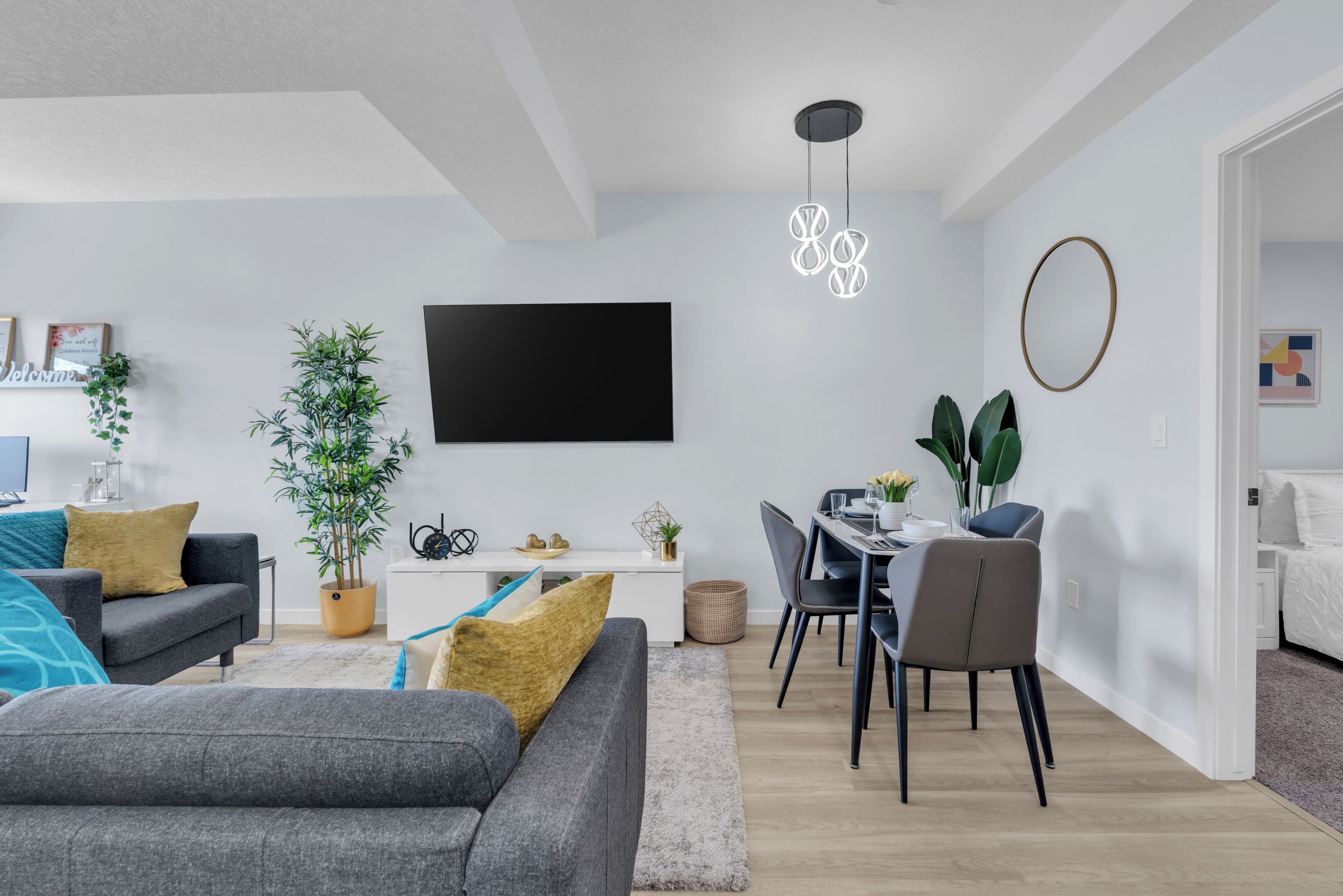 A modern living room with a grey sofa, a dining table with chairs, a flat-screen TV mounted on the wall, and a potted plant.