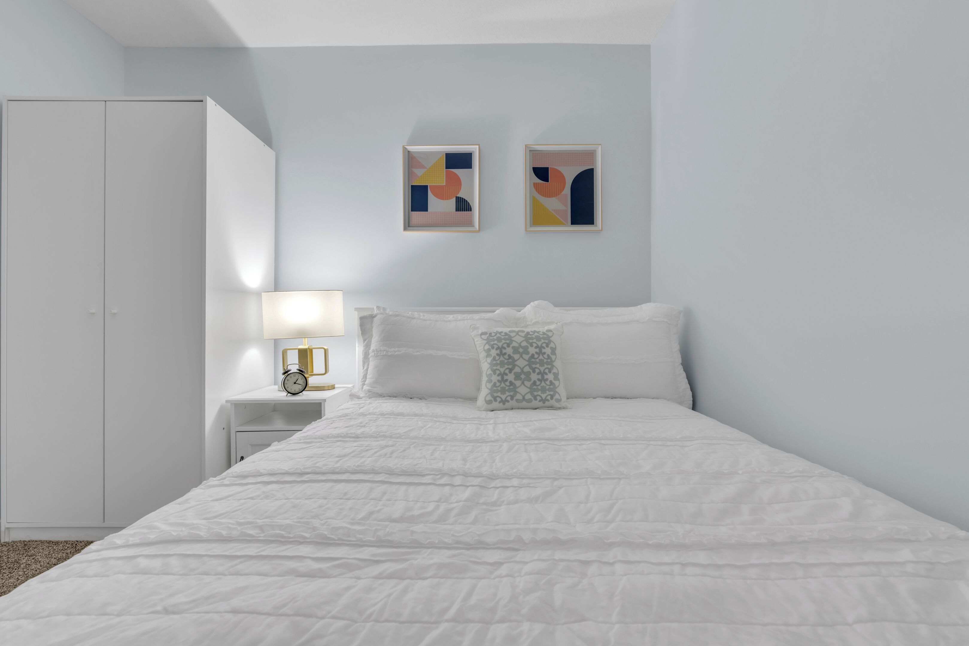 A bedroom with a large bed, a nightstand with a lamp, two framed artworks, and a wardrobe.