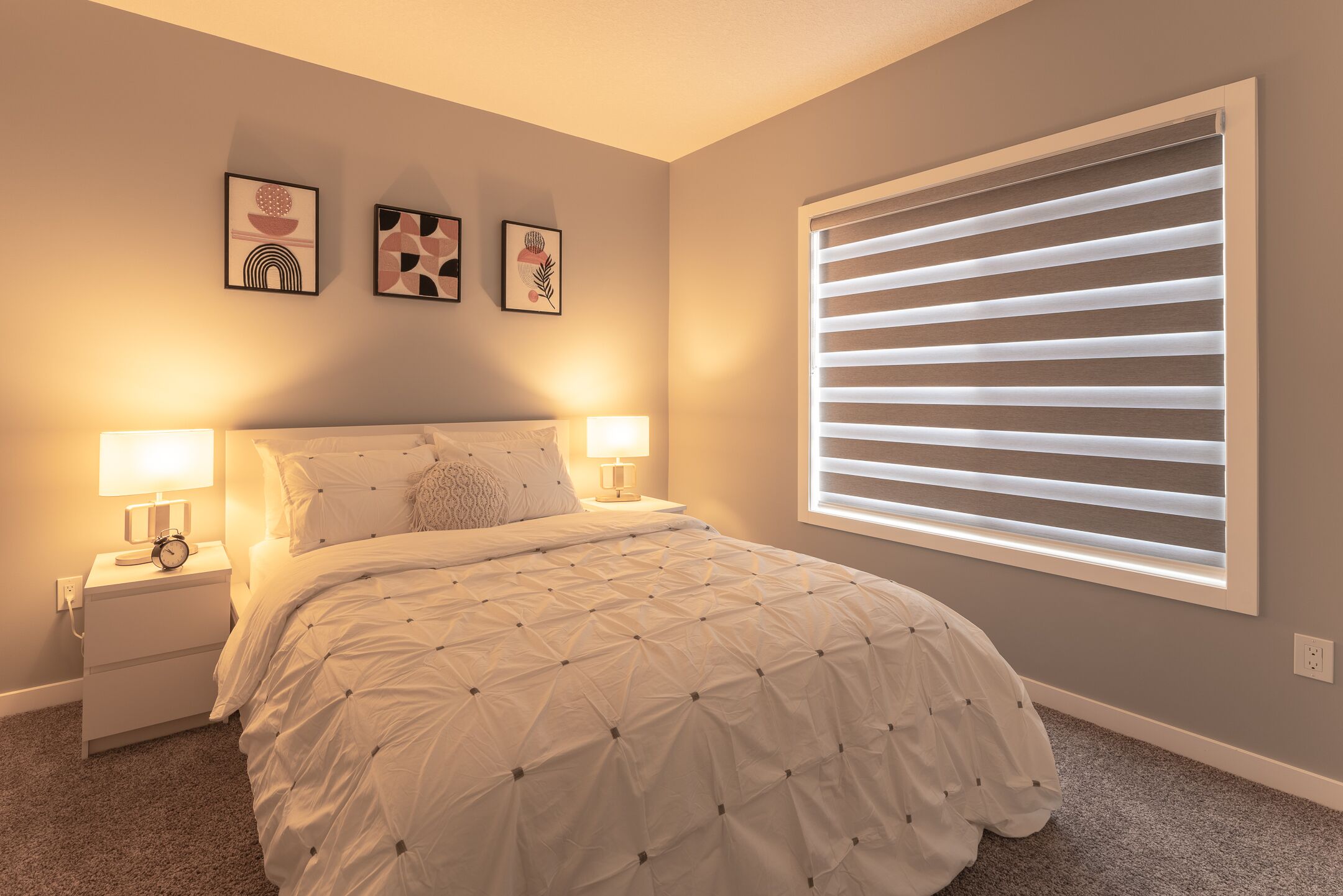Een slaapkamer met een bed, twee nachtkastjes met lampen, een raam met jaloezieën en drie decoratieve wandobjecten.