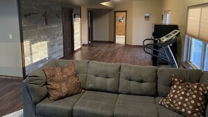 Ein geräumiges Wohnzimmer mit einem grauen Ecksofa, einem Laufband und einem großen Fenster.