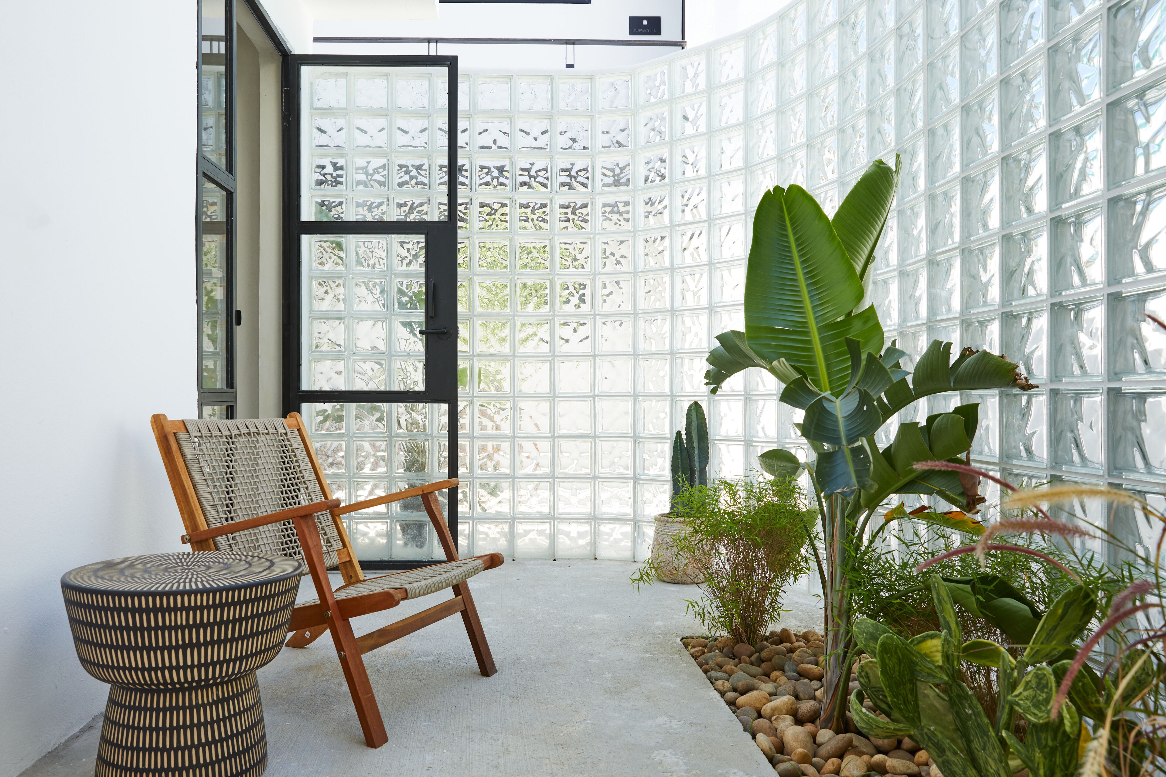 Um quarto com uma janela grande, uma cadeira de madeira, um banquinho de vime e diversas plantas.