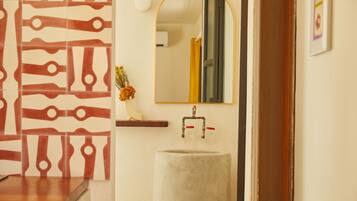 A bathroom with a patterned wall, a round sink, a mirror, and a wooden shelf with a vase.