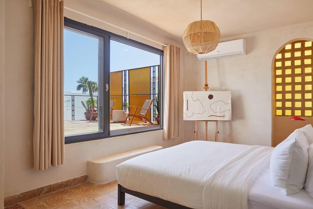 Une chambre d’hôtel avec un grand lit, un banc, une vue sur l’extérieur, un appareil de climatisation et une œuvre d’art accrochée au mur.