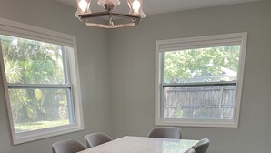 Comedor con mesa de mármol y sillas grises, ventanas amplias con vistas a árboles y suelo de madera.