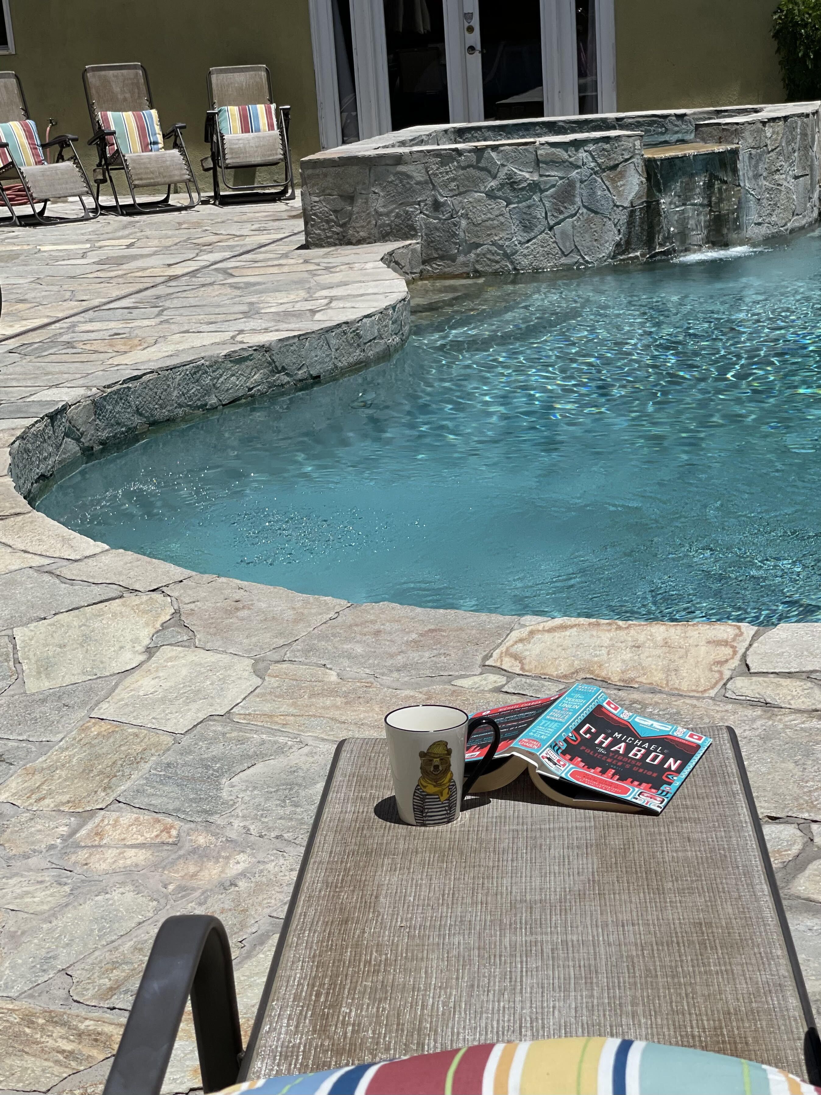 Un área de piscina con baldosas de piedra, sillas de descanso y un libro sobre una mesa.