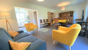 Una sala moderna con un sofá azul, sillones amarillos, una mesa de centro de vidrio y un comedor con sillas y mesa de madera.