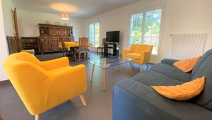 Una sala de estar con un sofá, sillones amarillos, una mesa de centro de vidrio, un televisor y una zona de comedor con un aparador y una mesa de madera.
