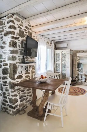 Una pared de piedra, una mesa de madera, sillas blancas y un televisor en una habitación con alfombra estampada.