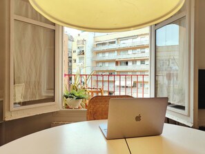 A room with a laptop on a table, a potted plant, and a view of a balcony with a railing.