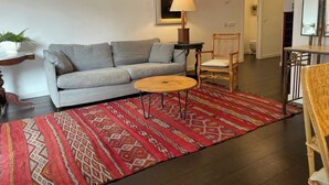 Un salon avec un canapé gris, une table basse ronde, un tapis à motifs, une chaise en bois et une petite table avec une lampe.