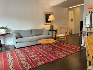 Un salon avec un canapé gris, une table basse ronde, un tapis à motifs, une chaise en bois et une petite table avec une lampe.