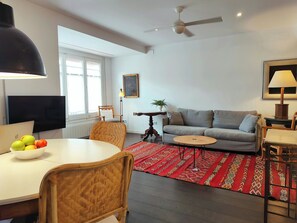 A modern living room with a sofa, a dining table with fruit, a TV, a ceiling fan, and a patterned rug. - F3- Spacious 2 bedroom. Sarrià - Hospital Clinic (Barcelona)
