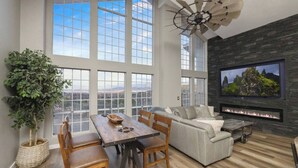 A spacious living room with a large window, a fireplace, a ceiling fan, and a television. - Lynx Lake Luxe Vacation Rental Wrapped in Modern Elegance | 6-BR House , Prescott | Sleeps 22 • Hot Tub • Game Rm • EV Charger • Horse Stalls (Prescott)