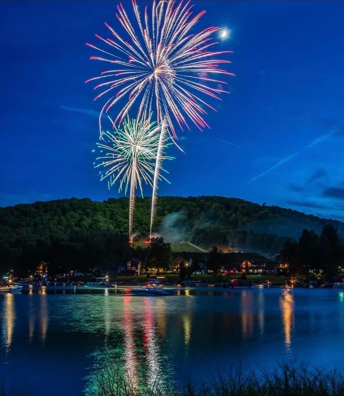 A nighttime view of a lake with fireworks reflecting on the water, surrounded by a forested hillside and buildings.