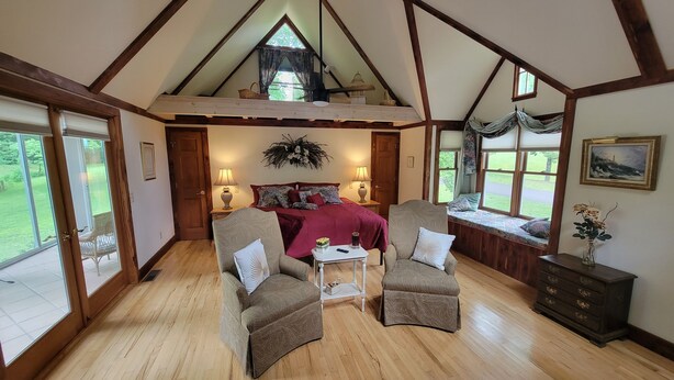 Une chambre avec un plafond en pente, un lit recouvert d’une couverture rouge, deux fauteuils, une petite table, une commode et un coin fenêtre d’où l’on peut admirer le paysage.