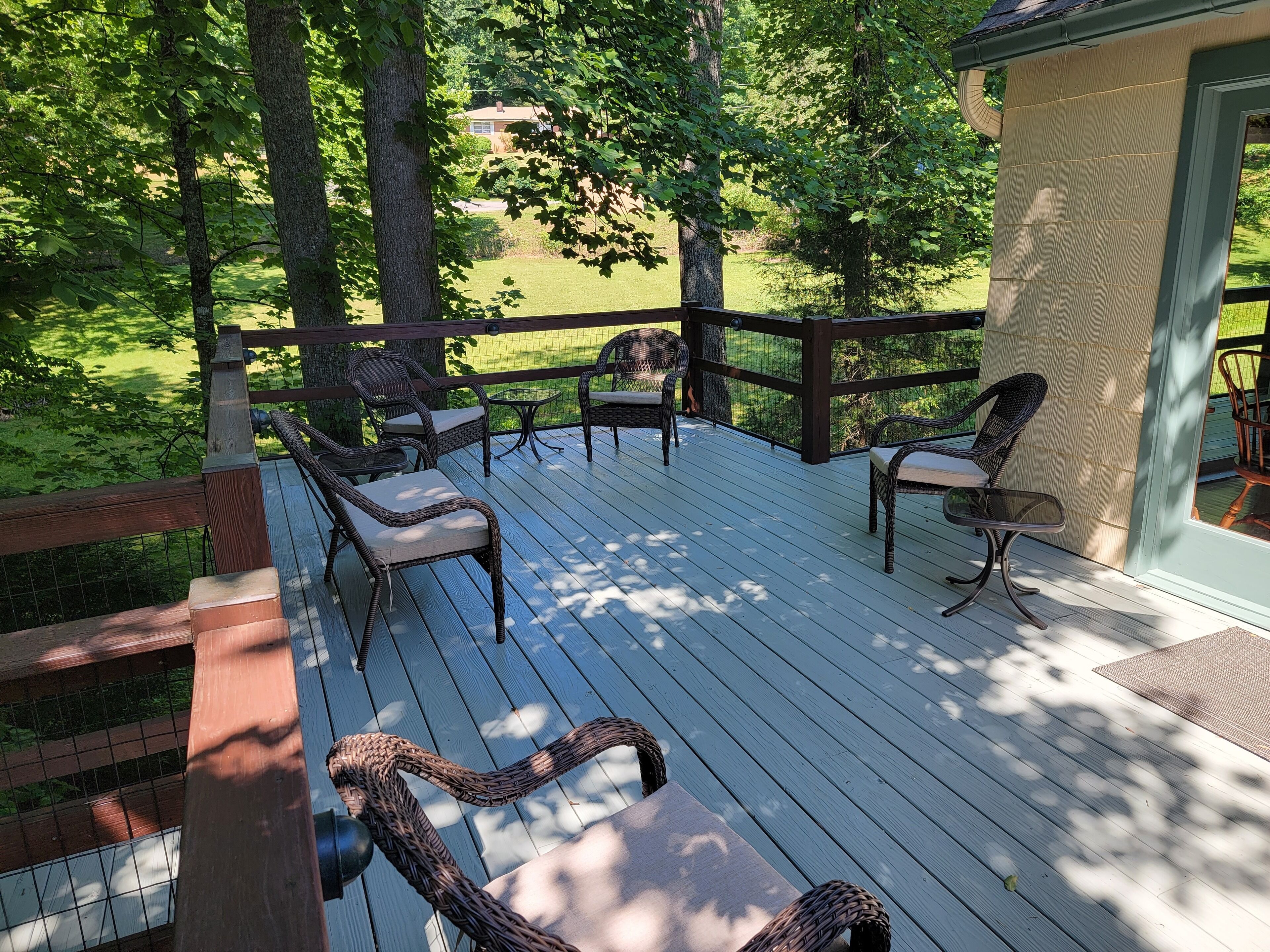 A wooden deck with wicker chairs and a small table, surrounded by trees and a house in the background.