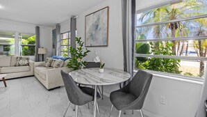 A modern living room with a marble coffee table, grey upholstered chairs, a beige sofa with patterned cushions, a large abstract painting, and a view of palm trees through large windows.