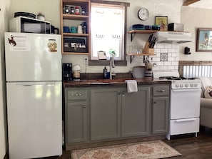 A kitchen with a refrigerator, stove, and cabinets. There is a clock on the wall and a window above the sink.