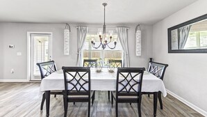A dining room with a white table, four chairs, a chandelier, and a view through the window. - Modern & Cozy ☀ Close to Beach ☀ 4 Bedrooms ☀ 2 Full Baths (Foley)