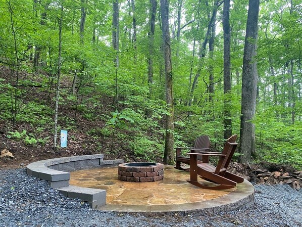 A circular stone fire pit with a wooden Adirondack chair and a bench in a forest setting.