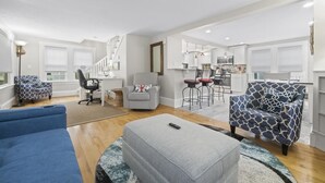 Een ruime woonkamer met een blauwe bank, een grijze poef en gestreepte stoelen. In de achtergrond is een keuken met moderne apparatuur en een bar met barkrukken te zien.