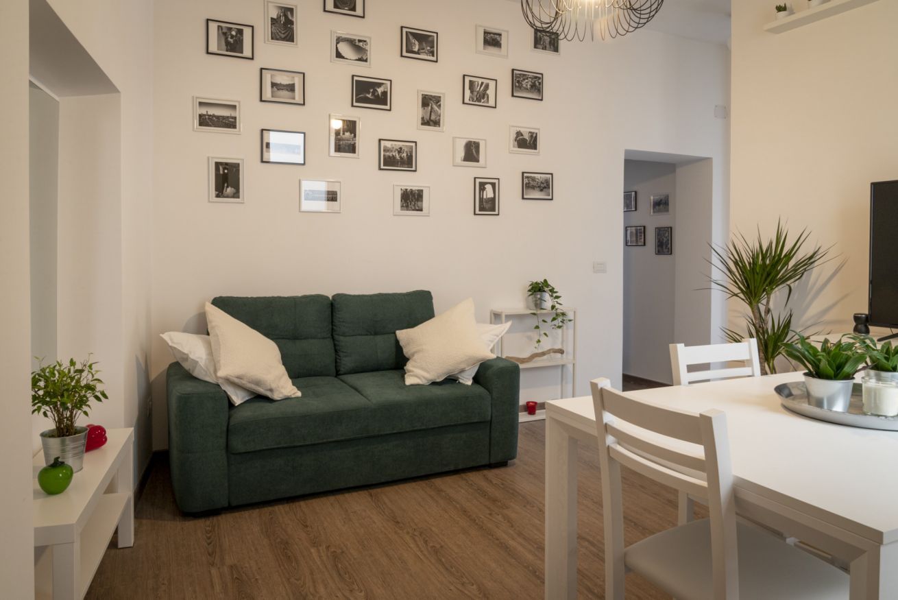 A modern living room with a green sofa, white cushions, a dining table with chairs, and a wall decorated with framed pictures.