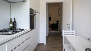 Eine schmale Küche mit Gasherd, Backofen und weißen Schränken. Im Vordergrund ist ein Esstisch mit einer Bank zu sehen.