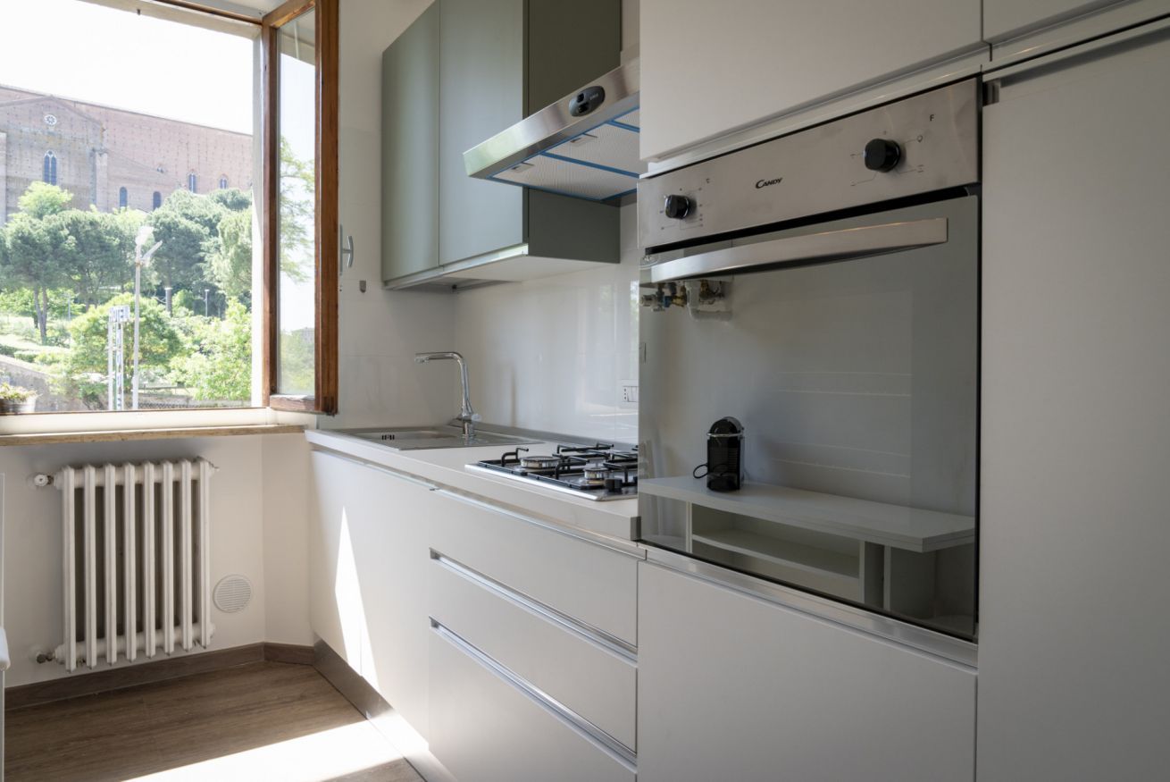Een moderne keuken met witte kasten, een inbouwoven en een raam met uitzicht op een bakstenen gebouw en groen.