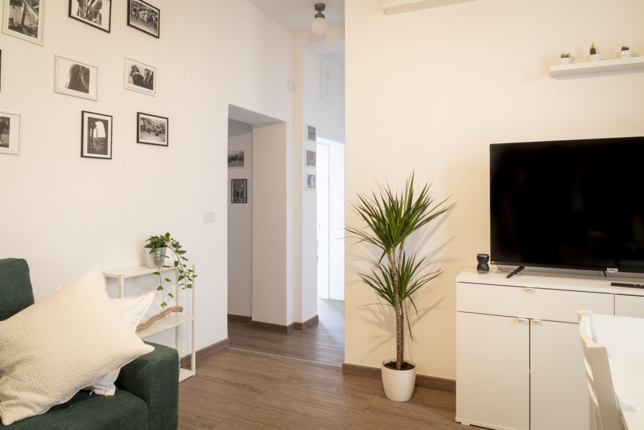 Een moderne woonkamer met een groene bank, een flatscreen televisie, een kamerplant en een gang die naar een andere kamer leidt.