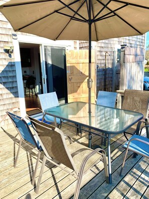 Eine Terrasse im Freien mit einem Glastisch, Metallstühlen, einem Holzzaun und einer Glasschiebetür, die zu einem Innenbereich führt.
