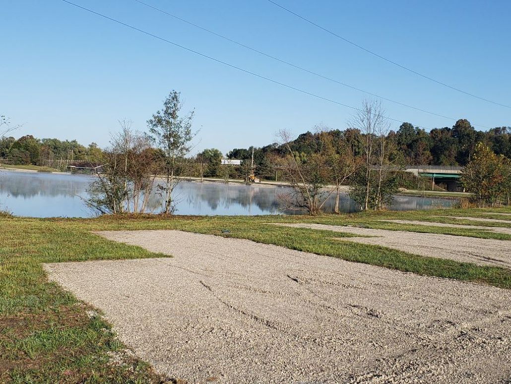 Un chemin de gravier mène à un plan d’eau, entouré d’arbres et avec un bâtiment visible au loin.