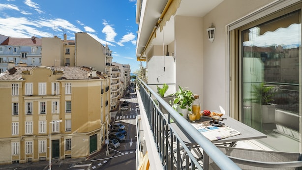 A balcony with a view of a city street, a table with a plant and a bottle, and a window with a curtain. - Downtown Cannes, Modern 1-Br. Apartment (Cannes)