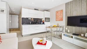 A modern living room with a white sofa, a dining table with chairs, a kitchen area with white cabinets, and a flat-screen TV mounted on a wooden wall. - Downtown Cannes, Modern 1-Br. Apartment (Cannes)