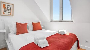 A modern hotel room with a bed, white bedding, red accents, a window, and a framed artwork on the wall.